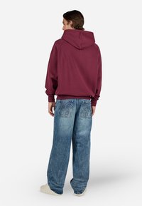 Man facing away wearing a maroon hoodie, loose blue jeans with stitched designs on back pockets, and white sneakers on a white background.