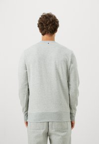 En krøllet hårperson iført en lys grå sweatshirt og matchende joggebukser står vendt bort fra en ensfarget hvit bakgrunn.