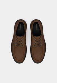 Smeđe cipele od brušene kože s okruglim vrhom, dizajnom s dvije rupice za vezanje i crnim gumiranim potplatom. Imaju glatku teksturu i detalje šavova.