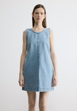Młoda kobieta stojąca twarzą do przodu, ubrana w bezrękawową, jasnoniebieską sukienkę dżinsową na jednolitym białym tle.