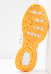 Semelle en caoutchouc orange vif avec des crampons texturés et des inserts blancs sur une semelle de chaussure de sport blanche, inclinée sur un fond blanc.