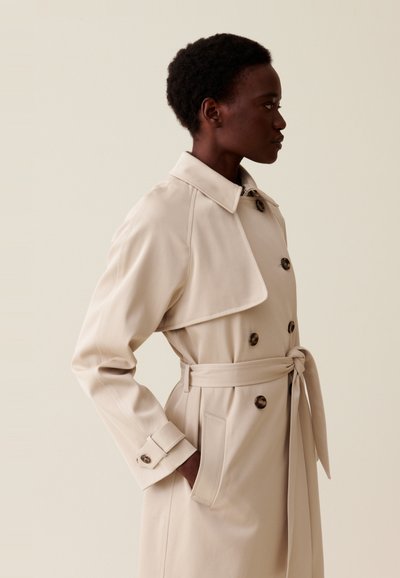 Trench coat beige con colletto classico, bottoni doppi, vita con cintura e tasche laterali; tessuto liscio con vestibilità sartoriale.