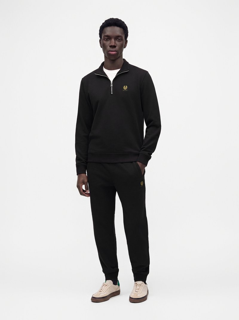 Homme portant un sweat zippé noir et un jogging noir assorti avec des baskets beige, debout devant un fond blanc uni.