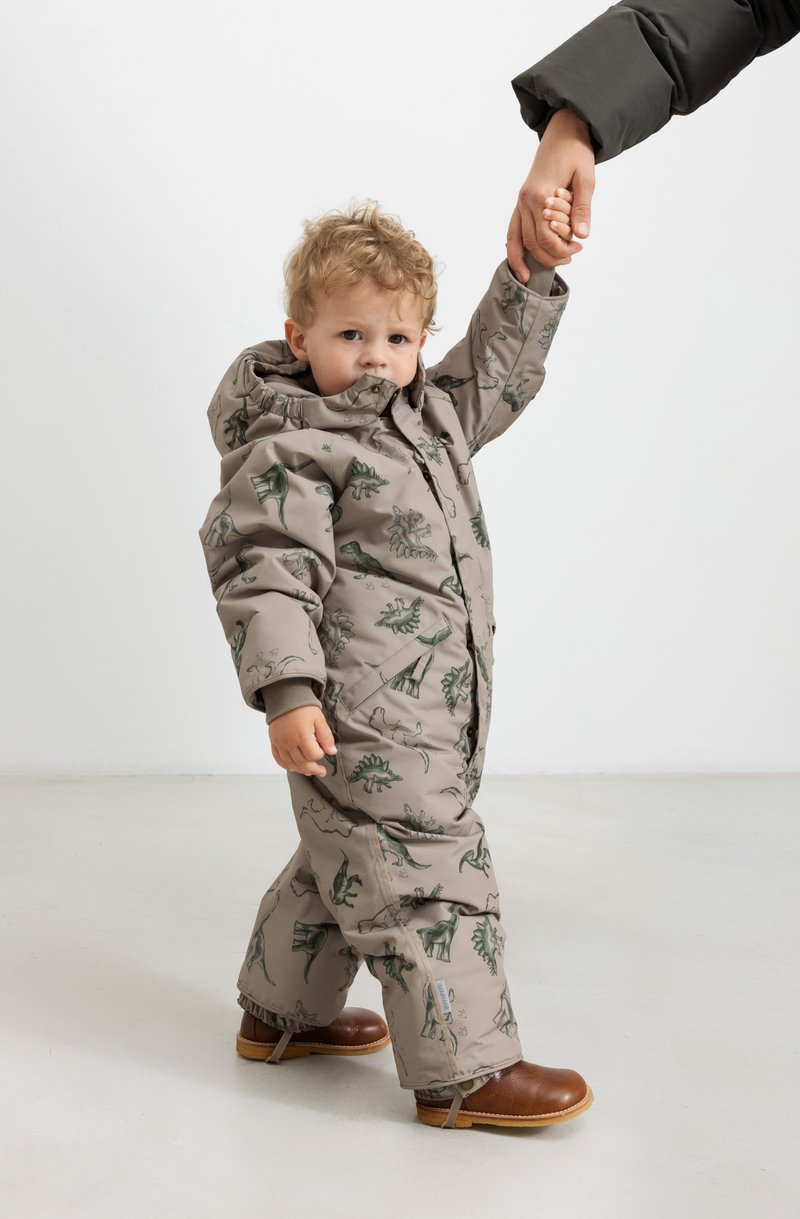 Pehmev, beež snowsuit roheline dinosauruste mustriga, kinnitatud kapuuts ja elastsed mansetid, kombineeritud pruunide saapadega. Laps hoiab täiskasvanu kätte.