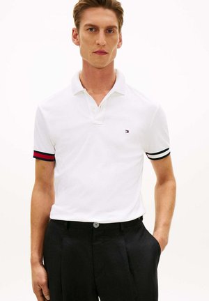 Giovane uomo che indossa una polo bianca con polsini delle maniche a strisce rosse e nere e pantaloni neri, in piedi con una mano in tasca.