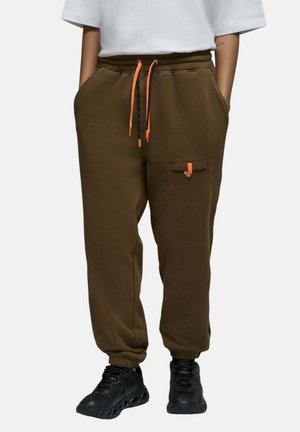 Personne portant un pantalon de jogging marron avec une taille élastique et des cordons orange, associé à des chaussures de sport noires et un haut blanc.