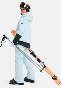 Jasnoniebieska kurtka narciarska z czarnymi akcentami, w zestawie z pasującymi spodniami. Narty i kijki w ręku, noszący czarny kask i przyciemnione gogle.