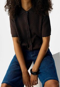 Haut marron en maille style polo à manches courtes, avec un motif texturé et des boutons. Assorti à un short en jean bleu et un bracelet noir.