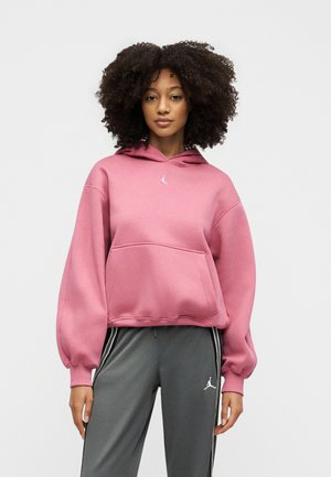 Mujer con cabello rizado vistiendo una sudadera rosa con capucha y pantalones deportivos grises con logotipos blancos de Jordan, de pie frente a un fondo liso.