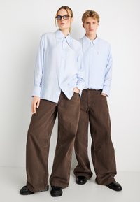 Ανοιχτό μπλε πουκάμισα με μεγάλα γιακά, συνδυασμένα με άνετα καφέ παντελόνια. Υφασμάτινη υφή με μαύρα παπούτσια δημιουργεί αντίθεση.