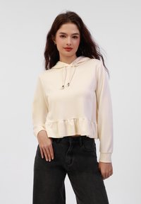 Felpa corta di colore crema realizzata in un tessuto morbido, con orlo a volant, maniche lunghe e lacci. Abbinata a jeans neri a vita alta.