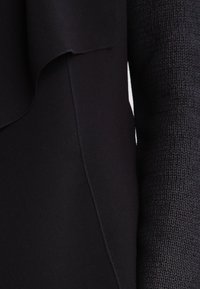 Czarny wielotexturowy element garderoby, łączący gładką, drapowaną tkaninę z grubszą, prążkowaną teksturą. Widoczny detal szwu.