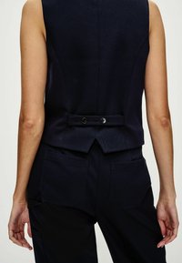 Marineblauw, op maat gemaakt vest met een gestructureerde stof, voorzien van een achtersplit en twee knopen. Gecombineerd met bijpassende broek met zakken aan de zijkant.