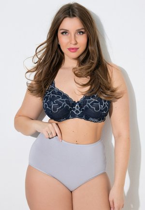 Frau mit langen braunen Haaren, die einen schwarzen Spitzen-BH und hellgraue hochtaillierte Slips trägt, steht vor einem weißen Hintergrund.