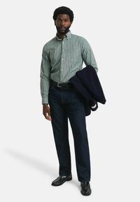 Groen en wit gestreepte overhemd met knopen, donkere spijkerbroek en zwarte schoenen; marineblauwe jas over de arm gehangen, staande pose tegen een witte achtergrond.