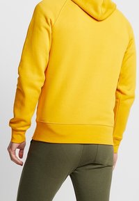 Sudadera con capucha amarilla de textura suave, con capucha y puños acanalados. Combinada con pantalones ajustados verde oscuro, mostrando un estilo casual.
