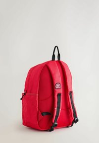Röd ryggsäck med en strukturerad yta, vadderade remmar, sidoficka och en cirkulär logotypapplikation. Har ett handtag för att bära ovanpå och ergonomisk design.