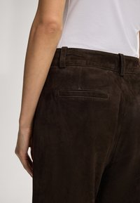 Braune Wildlederhosen mit glatter Textur, ausgestattet mit einer Gesäßtasche und einem Gürtelschlaufen. Die Ansicht zeigt die Hose von der Seite und von hinten.
