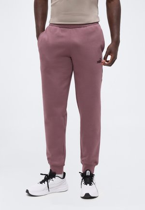 Mand iført lilla Puma joggingbukser med logo på låret og hvide Puma sneakers med sorte snørebånd står mod en ensfarvet baggrund.