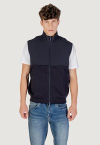 Gilet senza maniche con colletto alto, in tessuto blu navy, chiusura frontale con zip, due tasche laterali, abbinato a una t-shirt bianca sottostante e jeans blu.