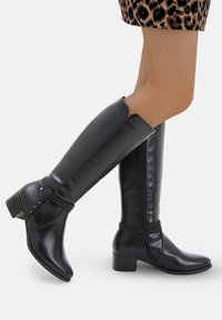 Botas negras hasta la rodilla con un acabado de cuero suave, cremallera lateral y un tacón bajo, con una correa decorada con tachuelas.