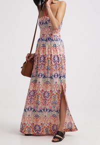 Femme portant une robe maxi sans manches colorée avec une fente sur le côté, des sandales noires et portant un sac à bandoulière marron.