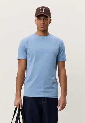 Ung mann med brun caps og lyseblå t-skjorte holder en svart bærepose mot en ensfarget lys bakgrunn.