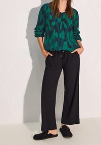 Zielona i czarna wzorzysta bluzka z długim rękawem o luźnym kroju, w połączeniu z czarnymi spodniami ze sznurkiem i czarnymi butami wsuwanymi.