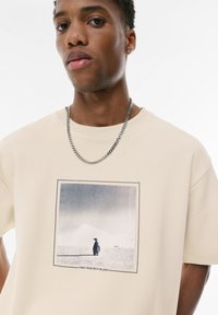 Beige kortærmet t-shirt med et grafisk print af en pingvin mod en lys baggrund. Har en rund halsudskæring og en sølvfarvet kæde.