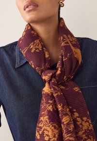Écharpe bordeaux à motif floral doré, en tissu doux, drapée autour du cou. Associée à une chemise en jean.