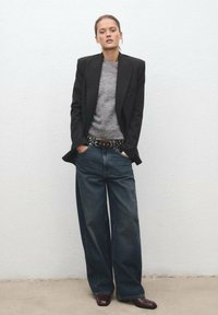 Črn oblikovan blazer čez sivo pulover, v kombinaciji s prostornimi temno modrimi džinsom in pasom s kovinskimi konicami. Model stoji z rokama v žepih.