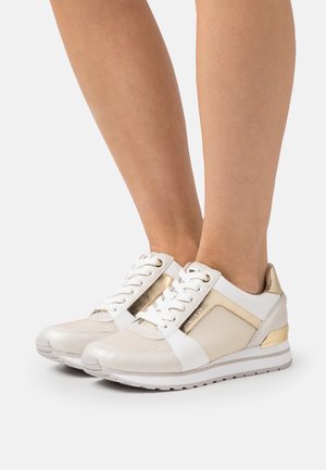 Baskets basses blanches et dorées portées aux pieds, avec des panneaux en mesh et cuir, des lacets blancs et des semelles en caoutchouc avec une légère semelle.