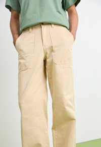 Beige cotton pants with a relaxed fit, featuring two front pockets and a subtle texture. Worn with a green short-sleeve shirt.