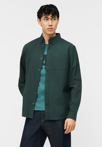 Νεαρός με κοντά σκούρα μαλλιά, φορώντας πράσινο σκούρο πουκάμισο με κουμπιά πάνω από μπλε-πράσινη μπλούζα με σχέδιο και σκούρο μπλε τζιν, κοιτάζει στο πλάι.