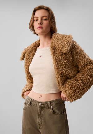 Ung kvinde iført en krøllet beige jakke over en beige crop top og højtaljede olivengrønne bukser, stående mod en ensfarvet baggrund.