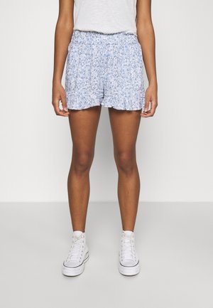Person, die weiße High-Top-Sneaker und hellblaue Shorts mit floralem Rüschensaum sowie ein weißes Oberteil trägt, steht vor einem einfarbigen Hintergrund.