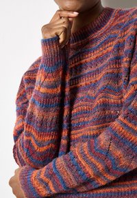 Knit sweater with zigzag pattern in orange, purple, and blue hues; features a textured fabric and ribbed cuffs with a round neckline.