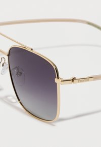 Kultaiset metalliset aurinkolasit, joissa on liukuväriset tummat linssit. Neliön muotoiset linssit ohuella ja tyylikkäällä kehykseellä sekä säädettävillä nenätyynyillä. Polaroid-logo näkyvissä.