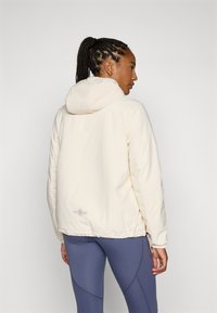 Lys beige hettejakke med en glatt tekstur, elastisk hem og subtil logo-detail på nedre rygg, kombinert med marineblå leggings.