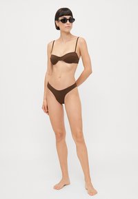 Brown bikini with a padded top featuring thin straps and a matching low-rise bottom. Model wearing black sunglasses, posing barefoot.