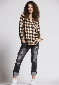 Bunt gemusterte Bluse mit schwarzen und goldenen ovalen Formen, kombiniert mit ausgewaschenen grauen Jeans und weißen Turnschuhen. Lockige Haare umrahmen das Gesicht.