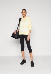 Haut à capuche tie-dye jaune et blanc avec des cordons, associé à un legging noir et des chaussures de sport noires. Le modèle tient un sac noir.