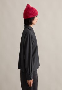 Svart langermet skjorte med spiss krage, kombinert med en teksturert, lys rød strikket lue. Sideprofil.