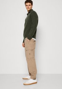 Khaki cargo kalhoty mají vícero kapes a texturovaný povrch, doplněné tmavě zeleným pleteným svetrem a bílými teniskami.