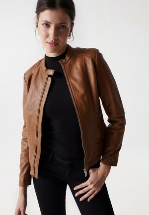 Veste en cuir marron au design ajusté, col rond et accents de zip argentés, associée à un haut noir à côtes et un jean sombre.