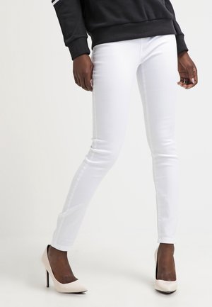 Slimfit witte broek gedragen door een staande persoon, gecombineerd met beige hoge hakken en een zwart top met lange mouwen.