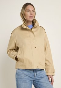 Beige cotton jacket with a high collar, snap buttons, and side pockets. Features shoulder epaulets and a relaxed fit. Worn over blue sweater.