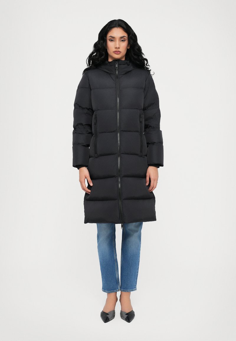 Black padded long coat with a zip closure, high collar, and side pockets. Features quilted design and wide sleeves. Worn over blue jeans.
