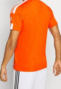 Oranžové sportovní tričko s bílými pruhy na ramenou, krátkými rukávy a hladkou texturou, nošené s bílými šortkami s černými pruhy.