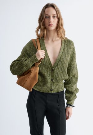 Jonge vrouw met een groene geribbelde cardigan, zwarte broek en een bruine suède schoudertas tegen een effen lichte achtergrond.